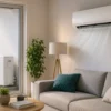Climatisation murale dans un salon moderne, unité extérieure visible par la fenêtre et canapé gris avec coussins, lampe sur pied et plante verte.